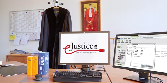 Das Bild zeigt einen Richterschreibtisch mit zwei Bildschirmen und davor Tastatur und Maus. Auf einem Bildschirm ist das eJustice-Logo zu sehen. Auf dem anderen Bildschirm die elektronische Akte.